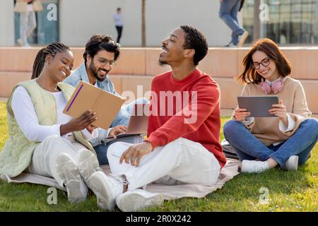Fröhliche internationale Millennials studieren, entspannen, auf dem Gras sitzen, Online-Unterricht auf einem Tablet ansehen Stockfoto