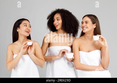 Drei verschiedene junge Frauen, die kosmetische, natürliche Gesichtscreme-Gläser halten, sich um ihre Haut kümmern, einen Spa-Tag genießen Stockfoto