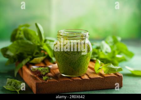 Hausgemachte Pesto-Sauce aus frischem Basilikum, Parmesan und Pinienkerne in Glasgefäß mit Löffel auf Holzbrett. Vorderansicht, Kopierbereich Stockfoto