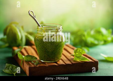 Hausgemachte Pesto-Sauce aus frischem Basilikum, Parmesan und Pinienkerne in Glasgefäß mit Löffel auf Holzbrett. Vorderansicht, Kopierbereich Stockfoto