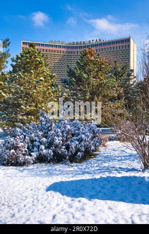 Blick auf das berühmte Hotel Usbekistan aus der Sowjetzeit, das teilweise hinter schneebedeckten Bäumen und Büschen versteckt ist. Gleich nach einem Wintersturm hinterließ ein weißes Licht Stockfoto