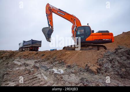 USt-Luga, Oblast Leningrad, Russland - 16. November 2021: Bagger Doosan lädt Lkw auf die Baustelle. Regnerischer Tag, dreckiger Boden. Stockfoto