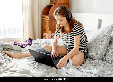 Hübsches Mädchen mit Kopfhörern und Laptop im Bett während des Webinars zum Online-Lernen. Ein wunderschöner Teenager lernt aus der Ferne mit einem Computer im Schlafzimmer Stockfoto