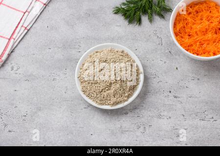 Schüsseln mit gemahlenen Sonnenblumenkernen, Karotten und einem Haufen frischen Dill auf grauem, strukturiertem Hintergrund, Draufsicht. Stufe der Zubereitung köstlicher veganer Schnitzel Stockfoto