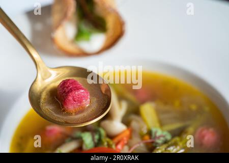 Selektiver Fokus einer rosafarbenen Gnocchi (Rote Bete) mit Gemüsesuppe auf einem goldenen Löffel. Spargel und Pilze in der Suppe. Weiße Platte. Stockfoto