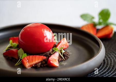 Selektivfokus eines kugelförmigen Bruchkuchens mit Erdbeeren und Brownie. Dessert mit glatten Oberflächen und Spiegelglasur. Rotes Dessert auf dem schwarzen Teller. Stockfoto