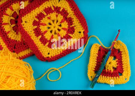 Oma-Quadrat in Gelb und Rot, gestrickt, mit zwei Oma-Quadraten und einer Kugel aus gelbem Garn teilweise sichtbar, alle auf einem aquablauen Hintergrund. Stockfoto