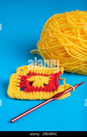 Granny Square in Gelb und Rot mit gelbem Garnknäuel und dunkelrotem Häkelhaken auf blauem Kingfisher-Hintergrund. Stockfoto