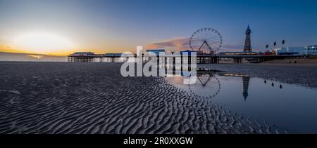 Ein Panoramablick auf den Blackpool Beach bei Sonnenuntergang mit Reflektionen des berühmten Turms und des Riesenrads am Central Pier auf dem nassen Sand. Stockfoto