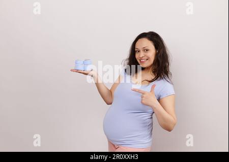 Lächelnde junge Schwangere zeigt mit dem Finger auf blaue gestrickte Babyschühchen auf ihrer Hand, lächelnd, isolierter weißer Hintergrund Stockfoto