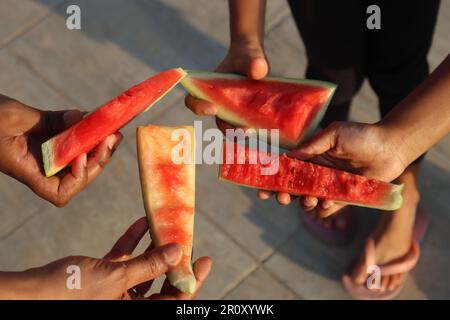 Wassermelonenscheiben mit Bissen, die an einem Sommertag in den kleinen Händen gehalten werden, mit Betonboden-Hintergrund. Sommerkonzept Stockfoto