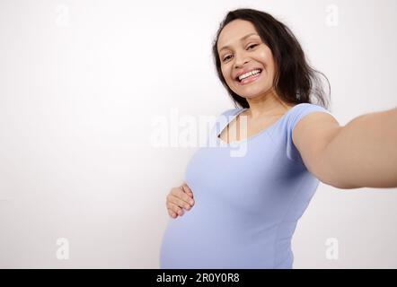 Frontaufnahme einer schwangeren Frau, die ihr Smartphone in der ausgestreckten Hand hält, lächelt und Selfie auf weißem, isoliertem Hintergrund macht Stockfoto