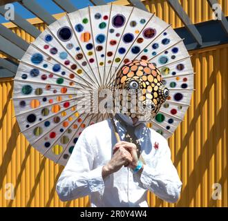 Ein Mann mit einer Meereskreatur-Maske, der einen Regenschirm mit farbigen ausgeschnittenen Kreisen hält, auf dem Riegelmann Boardwalk während der Coney Island Meerjungfrauen Parade. Stockfoto