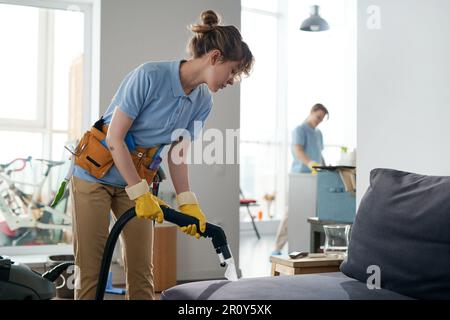 Reinigungskräfte arbeiten in der Wohnung im Team, saugen Möbel und reinigen das Zimmer Stockfoto
