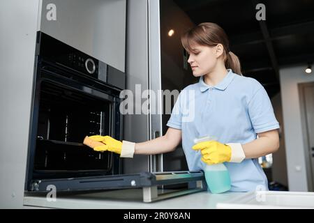 Junge Frau reinigt den Ofen mit Waschmittel in der Küche während der professionellen Hausarbeit Stockfoto