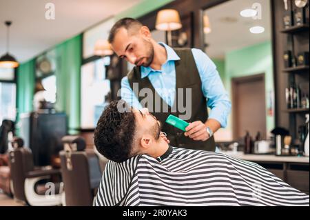 Männlicher Barbier mit Kammschnitt mit Schere des Kunden in gestreiftem Umhang während des Haarschnitts in einem modernen Friseursalon Stockfoto