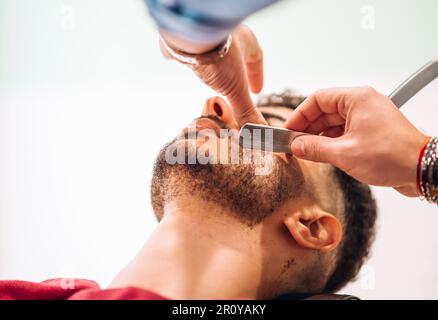 Unerkennbarer Barbier schneidet Bart eines ruhigen männlichen Klienten mit scharfem Rasierer in einem Friseursalon Stockfoto