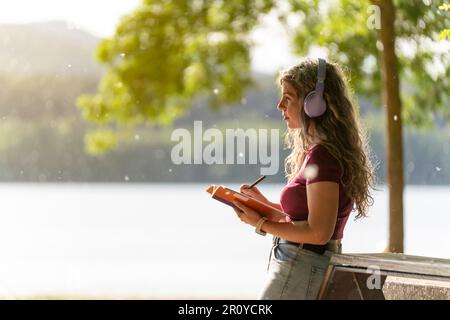 Eine Frau schreibt und hört Musik in der Nähe eines Sees in einem Park Stockfoto