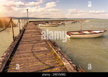 Hölzerner Pier in Balatonlelle mit bunten Booten auf dem blauen Himmel und dem Wasser des Balaton. Stockfoto