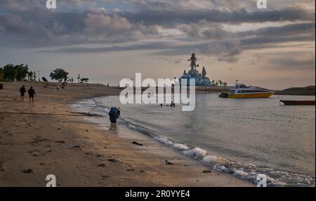 Pantai Jerman (deutscher Strand) in Kuta, Bali Indonesien bei Sonnenuntergang mit der Statue von Varuna (der Gott des Himmels, des Wassers und des Ozeans), während Menschen zu Fuß unterwegs sind Stockfoto