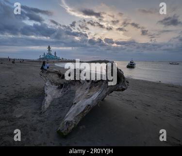 Pantai Jerman (deutscher Strand) in Kuta, Bali Indonesien bei Sonnenuntergang mit der Statue von Varuna (der Gott des Himmels, des Wassers und des Ozeans), während Menschen zu Fuß unterwegs sind Stockfoto