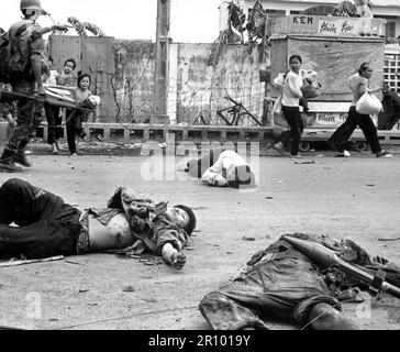 Mit Angst und Besorgnis in ihren Gesichtern und auf Drängen der südvietnamesischen Truppen schleicht sich Frauen und Kinder, die mit geborgenen Besitztümern beladen sind, an den Leichen von drei Vietcong vorbei, die in den Kämpfen getötet wurden. Mai 1968. Stockfoto