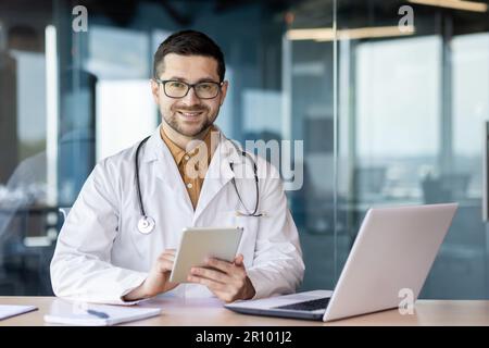 Porträt eines jungen Arztes. Eine Krankenschwester, eine medizinische Assistentin, die an einem Tisch in einer Krankenhauspraxis sitzt, an einem Laptop und einem Tablet arbeitet, lächelt und für die Kamera posiert. Stockfoto