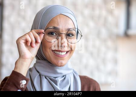 Nahaufnahme. Porträt einer jungen, wunderschönen muslimischen Frau in einem Hidschab, die lächelt, in die Kamera schaut und ihre Brille mit der Hand anpasst. Stockfoto