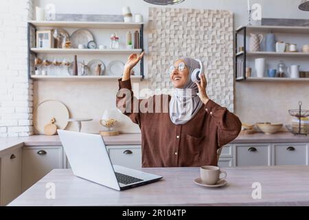 Eine junge Frau in einem Hidschab orientalischer Herkunft sitzt zu Hause in der Küche und hört Musik über Kopfhörer. Er hebt die Hände, tanzt, ruht sich aus, entspannt sich. Stockfoto