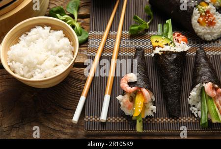 Bambusmatte mit leckeren Sushi-Zapfen, Essstäbchen und gekochtem Reis auf Holzhintergrund, Nahaufnahme Stockfoto