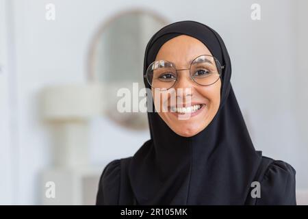 Nahaufnahme. Porträt einer jungen, wunderschönen muslimischen Frau in einem schwarzen Hidschab und einer Brille, die selbstbewusst in die Kamera lächelt. Stockfoto