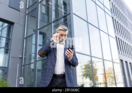 Ein besorgter älterer Mann in einem Anzug steht vor einem Bürozentrum und sieht enttäuschend auf den Telefonbildschirm und überrascht seine Brille mit der Hand auf dem Gesicht. Stockfoto