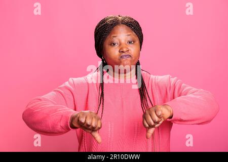 Enttäuschtes afroamerikanisches Modell zeigt Daumen-nach-unten-Symbol im Studio, macht negative Fehlschläge und Missbilligung mit Händen. Unzufriedene Frau, die Missbilligung und Ablehnung zeigt. Stockfoto