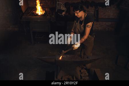 Nichts ist lohnender, als mit den Händen zu arbeiten. Eine junge Frau, die in einer Gießerei mit einem Hammer auf einen heißen Metallstab schlägt. Stockfoto