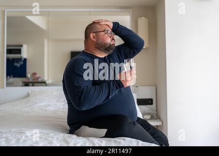 Trauriger, übergewichtiger, bärtiger Mann, der an starken Kopfschmerzen oder Migräne leidet, sitzt zu Hause im Bett. Stockfoto