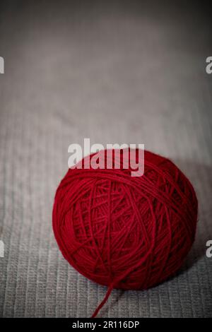 Rote Garnknäuel auf grauer gestrickter Decke mit Kopierbereich, Kunsthandwerk mit rotem Garn auf grauem gestricktem oder gestricktem Hintergrund. Kopierbereich, Stockfoto