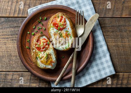 Backkartoffeln gefüllt mit Käse, Tomaten, grünen Zwiebeln und Eiern auf Keramikplatte auf Holzhintergrund, Draufsicht, Nahaufnahme Stockfoto