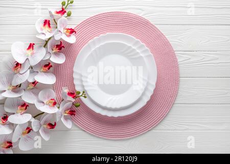 Festliches Ambiente mit pinkfarbenen Servietten und Orchideenblumen. Leere Teller auf weißem Holztisch. Draufsicht. Festliches Abendessen. Karte oder Menüvorlage, flach d Stockfoto