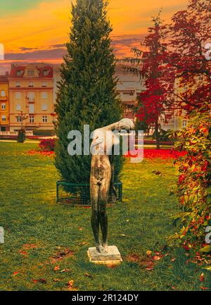 Eine idyllische Szene einer Bronzestatue in einem malerischen Park vor einem traditionellen Haus und Baum Stockfoto