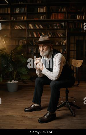 Eleganter älterer Geschäftsmann, der auf einem alten Sessel sitzt und eine Zigarre raucht Stockfoto