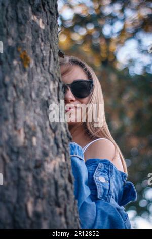 Eine junge Frau, die eine stilvolle Sonnenbrille und eine blaue Jeansjacke trägt und hinter einem Baumstamm in einer natürlichen Umgebung im Freien aus dem Fenster schaut Stockfoto