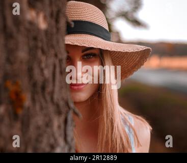 Eine junge Frau mit langen, goldblonden Haaren in braunem Hut hinter einem Baum Stockfoto