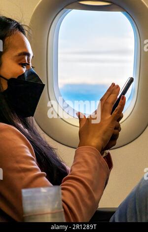 Asiatischer Passagier trägt Gesichtsmaske, Fensterplatz, mit Mobiltelefon auf dem Flug Stockfoto