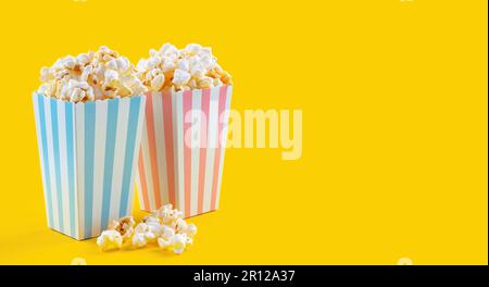 Zwei pinkfarbene und blau-weiß gestreifte Eimer mit leckerem Käsepopcorn, isoliert auf gelbem Hintergrund. Schachtel mit Streuung von Popcornkörnern. Schnell f Stockfoto