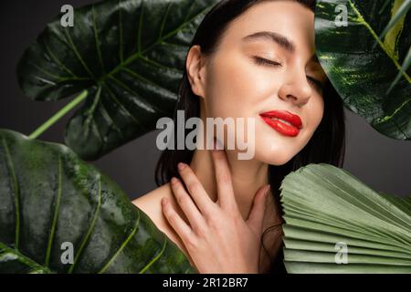 Zufriedene junge Frau mit braunem Haar und roten Lippen, die lächeln, während sie mit geschlossenen Augen neben tropischen grünen Palmenblättern mit Regentropfen posiert Stockfoto
