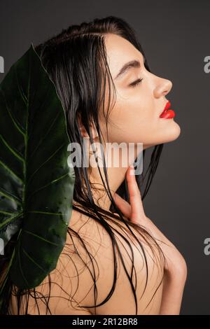 Seitenansicht einer wunderschönen jungen Frau mit braunem und nassem Haar, roten Lippen und perfekter Haut mit Hand am Hals neben verschwommener tropischer grüner Palme Stockfoto