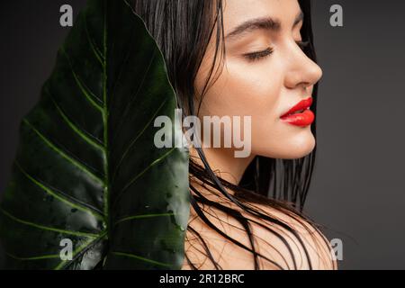 Charmante junge Frau mit braunem und nassem Haar, roten Lippen und perfekter Haut mit geschlossenen Augen neben verschwommenem tropischen grünen Palmenblatt isoliert Stockfoto