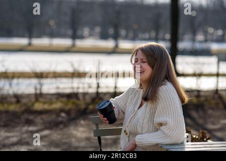 Lächelnde junge Frau in Strickpullover, die Kaffee im Straßencafé trinkt Stockfoto