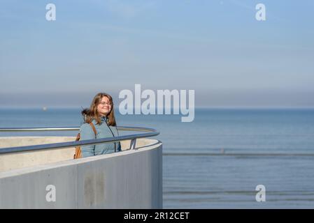 Lächelnde junge Frau im blauen Parka, die am sonnigen Frühlingstag in der Nähe des Meeres spazieren geht. Genießen Sie den Moment Stockfoto