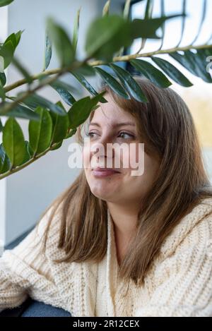 Lächelnde junge Frau in Strickpullover, die neben dem Fenster sitzt und Hauspflanzen hat Stockfoto